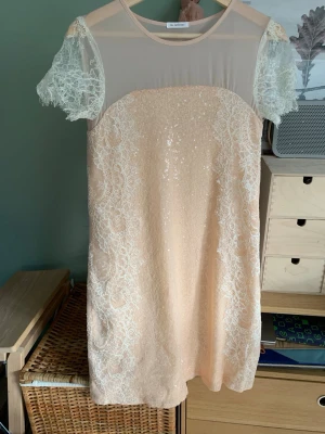 Champagnefärgad spetsklänning med paljetter 36 - Superfin kort klänning i champagnefärg. Klänningen har glittriga paljetter, spetsdetaljer och transparenta spetsärmar. Perfekt för dig som vill sticka ut med en elegant och feminin look.