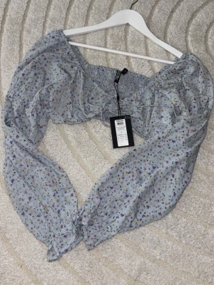 Blommig blus från Pieces med puffärm - Supersöt blommig blus från Pieces i ljusblått med små lila och gula blommor. Blusen har långa puffärmar, smock över bysten och en söt knytning framtill. Perfekt för vår och sommar, och ger en romantisk vibe till din outfit.