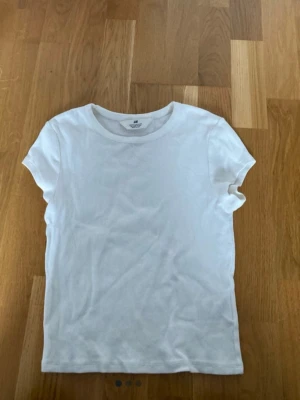 Vit ribbad t-shirt från H&M - En klassisk vit ribbad t-shirt från H&M med rund halsringning och korta ärmar. Perfekt basplagg i garderoben och enkel att matcha med allt. Tillverkad i mjuk bomull för en skön känsla.