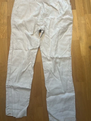 Beiga linnebyxor från H&M - Snygga och luftiga beiga linnebyxor med rak passform och resår i midjan. Byxorna har sidfickor och en bakficka, perfekta för en avslappnad stil. Tillverkade i ett lätt och svalt linnematerial som passar bra till varmare dagar.