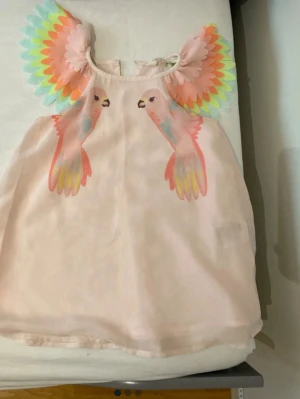 Pastellrosa klänning med papegojor - Superfin pastellrosa klänning med färgglada papegojmotiv framtill och 3D-detaljer som ser ut som vingar vid axlarna. Klänningen är ärmlös och har en lös passform, perfekt för dig som gillar unika och lekfulla plagg.