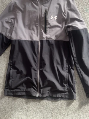 Grå och svart vindjacka Under Armour - Snygg vindjacka från Under Armour i tvåfärgad design med grå ovandel och svart nedtill. Jackan har hel dragkedja, bröstficka med dragkedja och huva. Perfekt för blåsiga dagar och sportiga outfits.