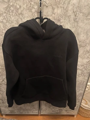 Svart hoodie med känguruficka - En svart hoodie med klassisk känguruficka och huva. Hoodien har en enkel och stilren design med ribbade muddar vid ärmslut och nederkant. Perfekt för en avslappnad och cool look. Materialet ser ut att vara mjukt och bekvämt, troligen bomullsblandning.