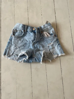 Ljusa denimshorts med slitningar - Säljer ett par ljusblå denimshorts med råa kanter och slitna detaljer framtill och baktill. Klassisk femficksmodell med knapp och dragkedja. Perfekta för varma dagar och ger en avslappnad vibe.