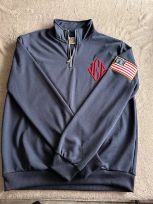 Jeffrey Epstein Tröja - Snygg mörkblå tröja med halv dragkedja och ståkrage. På bröstet finns ett rött broderat monogram och på ärmen sitter en patch med amerikanska flaggan. Tillverkad i mjukt material, perfekt för en avslappnad streetwear-look.