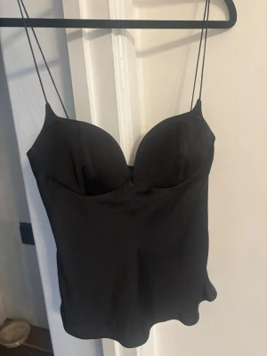 Svart satinlinne med tunna axelband från Zara - Stilrent svart linne från Zara  satin med tunna axelband och formade kupor. Linnet har en lätt glansig finish och en figurnära passform som ger en snygg siluett. Perfekt att styla med jeans eller kjol för en trendig look.