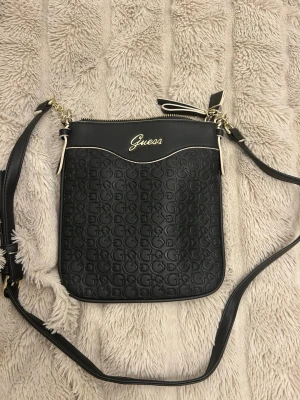 Svart axelväska från Guess - Snygg svart axelväska från Guess med präglat G-mönster och guldfärgade detaljer. Väskan har justerbar axelrem, dragkedja upptill och logotyp framtill. Insidan är fodrad med mönstrat tyg och har flera fack för småsaker. Aldrig använd.
