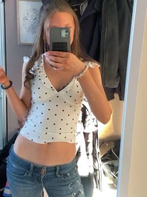 Vit prickig croppad topp med volang - Snygg vit croppad topp med svarta prickar och volangdetaljer vid axlarna. Djup v-ringning och figurnära passform som framhäver midjan. Perfekt för en trendig och ungdomlig stil. Använd några få gånger😊