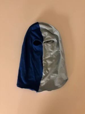 Tvåfärgad balaclava i blått och grått - Säljer en balaclava i mjukt tyg med tvåfärgad design, ena sidan är mörkblå och andra sidan ljusgrå. Modellen har ett ögonhål och är perfekt för att täcka hela huvudet. Materialet känns stretchigt och bekvämt.