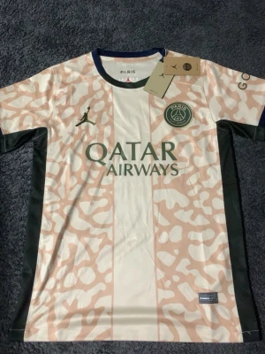 PSG x Jordan matchtröja rosa/vit - Säljer en Paris Saint-Germain matchtröja i samarbete med Jordan. Tröjan är rosa och vit med camo-mönster, gröna detaljer och PSG-logga på bröstet. Qatar Airways som sponsor framtill. Perfekt för fotboll och streetwear.