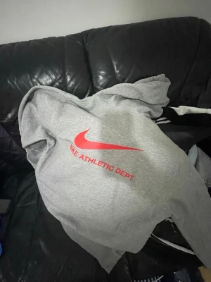 Grå Nike hoodie med röd logga - Snygg grå hoodie från Nike med stor röd Swoosh och texten 'NIKE ATHLETIC DEPT.' på bröstet. Hoodien har huva och tryck även på ena ärmen. Perfekt för en sportig och avslappnad stil.