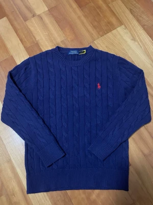 Mörkblå kabelstickad tröja Polo Ralph Lauren - Denna klassiska Polo Ralph Lauren kabelstickad tröja i marinblått har den ikoniska röda broderade Polo Pony på bröstet. Tillverkad av 100 % bomull, erbjuder den en tidlös design med en bekväm rund hals, perfekt för mångsidig styling. 
