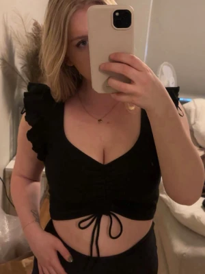 Svart croppad topp med volangärmar - Svart croppad topp från Zara med v-ringning, volangärmar och snörning framtill. Toppen är ribbad och har en tight passform som framhäver midjan. Perfekt för dig som gillar trendiga och feminina detaljer. Man kan dra åt snörerna hur ” liten ” man vill ha den. I storlek M men skulle säga mer som en S.