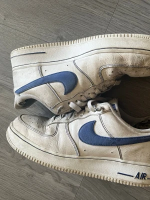 Nike Air Force 1 vita/blå sneakers - Klassiska Nike Air Force 1 sneakers i vitt läder med blå swoosh och detaljer. Lågt skaft och robust sula med AIR-logga på sidan. Perfekta för dig som gillar retrostil och streetwear. Ikonisk modell med tydlig Nike-branding på häl och plös.
