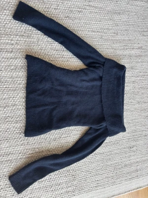 Mörkblå stickad of shoulders tröja från Ginatricot - Säljer en mörkblå stickad of shoulders med bred ribbad krage och långa ärmar. Tröjan är mjuk och har en smal passform, perfekt för kyliga dagar. Materialet känns ulligt och mysigt, och färgen är klassiskt marinblå.