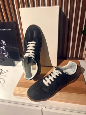 Maison margiela svarta - Stilrena svarta sneakers med vit sula och vita skosnören. Ovandelen är i mocka och läder, med klassisk låg profil och retrovibbar. Perfekta för dig som gillar minimalistisk och tidlös design.
