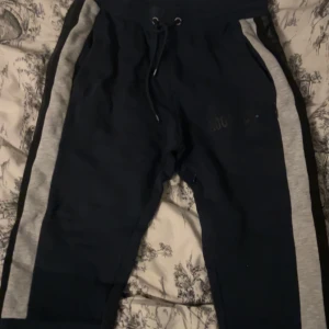 Mjukisbyxor från Hoodrich, blå/grå - Snygga mjukisbyxor från Hoodrich i mörkblått med grå sidopaneler och svarta detaljer. Byxorna har dragsko i midjan och är tillverkade i mjukt bomullsmaterial. Perfekta för chill och streetwear-stil.