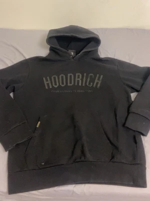 Svart hoodie från Hoodrich - Cool svart hoodie från Hoodrich med stor logga på bröstet och texten 'From Nothing To Something'. Klassisk passform med huva och känguruficka. Materialet är mjukt och bekvämt, perfekt för chill dagar. Metalltag med Hoodrich-logga vid fickan. Pris kan diskuteras, jag är öppen för att sänka så hör av er. 
