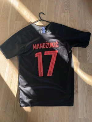 Svart fotbollströja Mandžukić 17 - Snygg svart fotbollströja med röd text och siffror på ryggen. Mandžukić och nummer 17 tryckt i rött. Kortärmad modell i lätt och ventilerande material, perfekt för träning eller match.