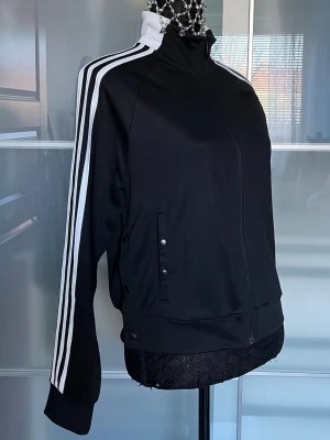Svart Adidas track jacket - Snygg svart track jacket från Adidas med klassiska vita ränder längs ärmarna och vit hög krage. Dragkedja framtill och tryckknappar vid fickorna. Adidas-logga i vitt på ryggen. Perfekt till träning eller streetwear.