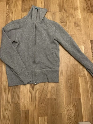 Grå zip tröja - Säljer en stilren grå hoodie med dragkedja framtill och två fickor. Tröjan har hög krage och ribbade muddar vid ärmslut och nederkant. Tillverkad i mjukt bomullsmaterial, perfekt för en avslappnad look.