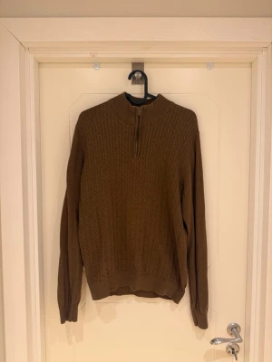 Brun stickad tröja med halv dragkedja - Säljer en brun stickad tröja med halv dragkedja och hög krage. Tröjan har ett diskret kabelstickat mönster framtill, långa ärmar och ribbade muddar vid ärmslut och nederkant. Perfekt för kyliga dagar och enkel att matcha.