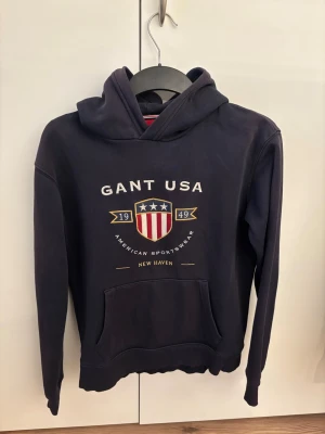 Mörkblå hoodie från GANT USA - Snygg mörkblå hoodie från GANT med tryck på bröstet där det står 'GANT USA' och ett sköldmärke med amerikanska flaggan. Hoodien har huva, känguruficka och ribbade muddar. Perfekt för dig som gillar sportig och klassisk stil. Materialet känns mjukt och bekvämt.