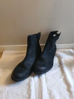 Svarta stövletter i skinn med dragkedjaå - Snygga svarta stövletter i skinn med rund tå och låg klack. Dragkedja på insidan och dekorativ knappdetalj vid ankeln. Klassisk och stilren modell som passar till många outfits.