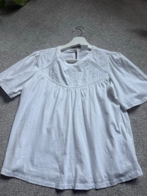 Vit blus med broderi från Zara - Söt vit blus från Zara med fina broderade detaljer upptill och rund halsringning. Blusen har korta ärmar och lätt utställd passform. Perfekt till jeans eller kjol för en fräsch look.