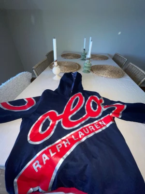 Mörkblå Ralph Lauren hoodie med tryck - Snygg mörkblå hoodie från Ralph Lauren med stort rött och vitt POLO-tryck över bröstet och ärmen. Huva med stjärnmönster i vitt på blå botten. Röd och vit rand vid nederkant och ärmslut. Perfekt statement-plagg för dig som gillar streetstyle.