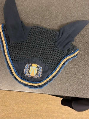 Blått virkat hästhuva - Säljer ett mörkblått virkat hästhuva med ljusblå kant och tygöron. Skyddet har två hål för öronen och är perfekt för att skydda hästens öron mot insekter. Passar till ridning och träning. Knappt använt. Storlek full.