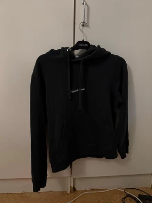 Jack and Jones hoodie  - Mörk grön hoodie från Jack and Jones med huva och dragsnören. Framtill finns en broderad text och en klassisk magficka. Hoodien har långa ärmar och är tillverkad i mjukt material, perfekt för en avslappnad stil.