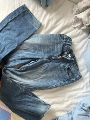 Blå raka jeans med slitningar - Ett par blå jeans med rak passform och tydliga slitningar samt ljusare tvätt på låren. Jeansen har klassisk femficksdesign och råa kanter vid bensluten. Perfekta för en avslappnad och trendig look.