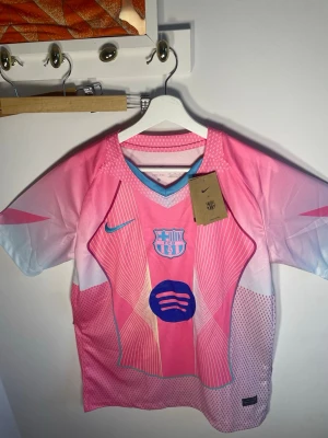 FC Barcelona matchtröja Nike - Snygg FC Barcelona matchtröja från Nike i rosa och ljusblått med grafiskt mönster och klubbmärke på bröstet. Tröjan har korta ärmar, V-ringning och är tillverkad i lätt polyester – perfekt för fotboll eller träning.