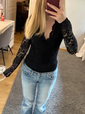 Svart spetsig långärmad body - Snygg svart långärmad body med spetsdetaljer och v-ringning. Toppen har figurnära passform och transparenta ärmar i spets, vilket ger en trendig och elegant look. Perfekt att styla med jeans eller kjol. Den är helt ny och säljes pga att den är för stor💗💗💗