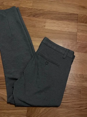 Mörkgrå kostymbyxor med smal passform - Snygga mörkgrå kostymbyxor med smal passform och klassiska pressveck. Byxorna har bälteshällor, bakficka med knapp och är tillverkade i ett mjukt tyg som ger en stilren look. Perfekta för dig som vill ha en clean och modern stil.