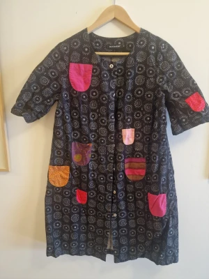 Mönstrad klänning med färgglada fickor - Glada Rocken, en klassisk klänning från Marimekko med svart bas och vitt cirkelmönster. Klänningen har flera färgglada fickor i rosa, orange och lila nyanser samt knappar framtill. Modellen är knälång med korta ärmar och tillverkad i bomull.