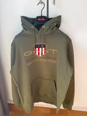 GANT Shield Hoodie - Snygg olivgrön hoodie från Gant med broderad logga och texten 'American Sportswear' på bröstet. Klassisk huva med dragsko och stor magficka framtill. Mjukt material, perfekt för en avslappnad stil. 🎉 OBS! Hoodien har 5 punktstorleks prickar som har framkommit från blekning vid okänt tillfälle, dock finns dem endast på kanten av ryggen och inte på armar eller framsida, därav pris även fast tröjan är som ny och typ aldrig använd! 🎉För mer info skriv dm! (Köpt för 1400 kronor på Boozt)