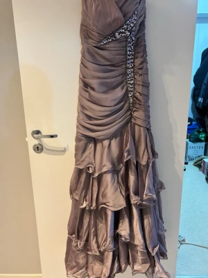 Lång lila balklänning med volanger - Supersnygg långklänning i lila satin med flera lager volanger och draperad överdel. Klänningen har en glittrig detalj framtill som ger extra wow-faktor. Perfekt för bal eller andra speciella tillfällen där du vill sticka ut. Finns en tillagd scarf!