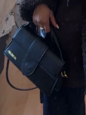 Svart handväska från Jacquemus - Stilren svart handväska från Jacquemus med guldiga detaljer och logotyp på framsidan. Väskan har en rektangulär form, bred lockstängning och justerbar axelrem. Tillverkad i slätt läder som ger en lyxig känsla och passar perfekt till trendiga outfits.