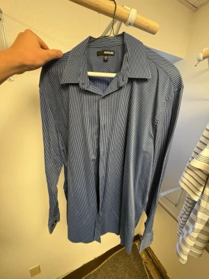 Blå randig skjorta från Murano - Säljer en blå långärmad skjorta från Murano med smala vita ränder. Skjortan har klassisk krage och knäppning framtill. Perfekt för dig som gillar stilrena och tidlösa plagg. Materialet är mjukt och känns bekvämt mot huden.