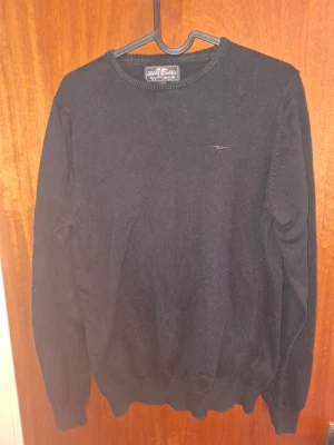 Svart stickad tröja från Hampton Republic - En klassisk svart stickad tröja från Hampton Republic med rund hals och diskret logga på bröstet. Tröjan har långa ärmar och ribbade muddar vid ärmslut och nederkant. Perfekt för lager-på-lager och enkel att matcha med jeans eller chinos.