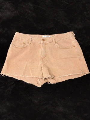 Beige manchester shorts med rå kant - Säljer ett par beige manchester shorts med rå, fransig kant och klassisk femficksdesign. Shortsen har hög midja och stängs med knapp och dragkedja. Perfekta för varma dagar och ger en avslappnad vibe.
