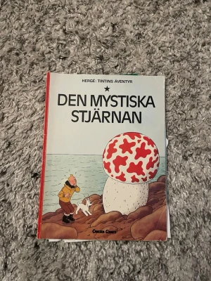 Den mystiska stjärnan Tintin-album - Säljer seriealbumet 'Den mystiska stjärnan' av Hergé ur Tintins äventyr. Omslaget är färgglatt med röd rygg och motiv av Tintin, Milou och en stor svamp. Perfekt för dig som gillar klassiska serier och äventyr.
