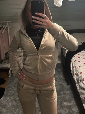 Juicy Couture Beige - Snygg beige hoodie i mjuk velour med dragkedja framtill och två stora fickor. Tröjan har huva med dragsko och ribbade muddar vid ärmslut och nederkant. Perfekt för en chill och avslappnad stil. J:et på dragkedja är lite söndrigt inget som påverkar funktion🩷💗😍🙏🏻