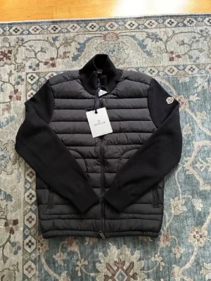 Moncler cardigan - Snygg svart cardigan från Moncler med quiltad front och stickade svarta ärmar. Västen har dragkedja framtill, två sidofickor med dragkedja och klassisk Moncler-logga på ärmen. Perfekt för lager-på-lager och streetstyle.