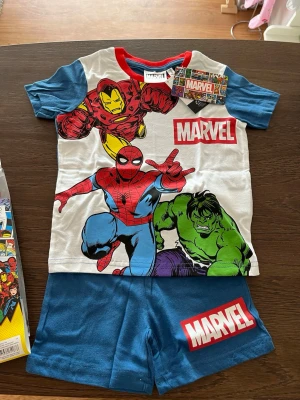 Marvel set med Spiderman, Iron Man & Hulk - Snyggt Marvel set med t-shirt och shorts. T-shirten har tryck av Spiderman, Iron Man och Hulk i färgglada toner samt Marvel-logga. Blå ärmar och röd krage. Matchande blå shorts med Marvel-logga på benet. Perfekt för alla superhjältefans!