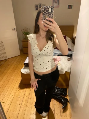 Vit prickig omlott topp med volang - Säljer en vit omlott topp med svarta prickar och volangdetaljer på axlarna. Toppen har v-ringning och är ärmlös, perfekt för dig som gillar en feminin och trendig stil. Passformen är tight och materialet känns mjukt och bekvämt.
