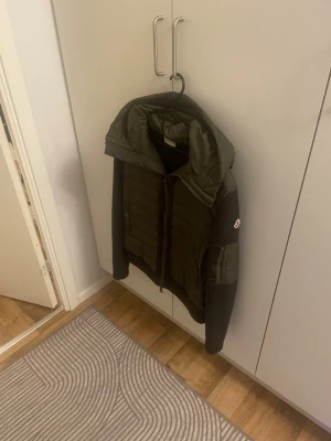 Grön pufferjacka från Moncler - Säljer en grön pufferjacka från Moncler med stickade ärmar och huva. Jackan har quiltad front, två fickor med knappar och en ärmficka med Moncler-logga. Perfekt för kalla dagar och snygg att matcha med jeans.