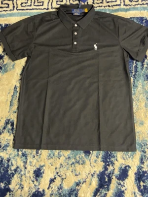 Svart pikétröja från Polo Ralph Lauren - Klassisk svart pikétröja från Polo Ralph Lauren med vit broderad logga på bröstet. Tröjan har korta ärmar, krage och knappar framtill. Perfekt för en stilren look och tillverkad i mjuk bomull.